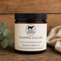 An amber glass jar of Black Diamond Tallow Co. Whipped Tallow with a black lid and cream label, styled on a rustic wooden surface with a eucalyptus sprig and a beige cloth.